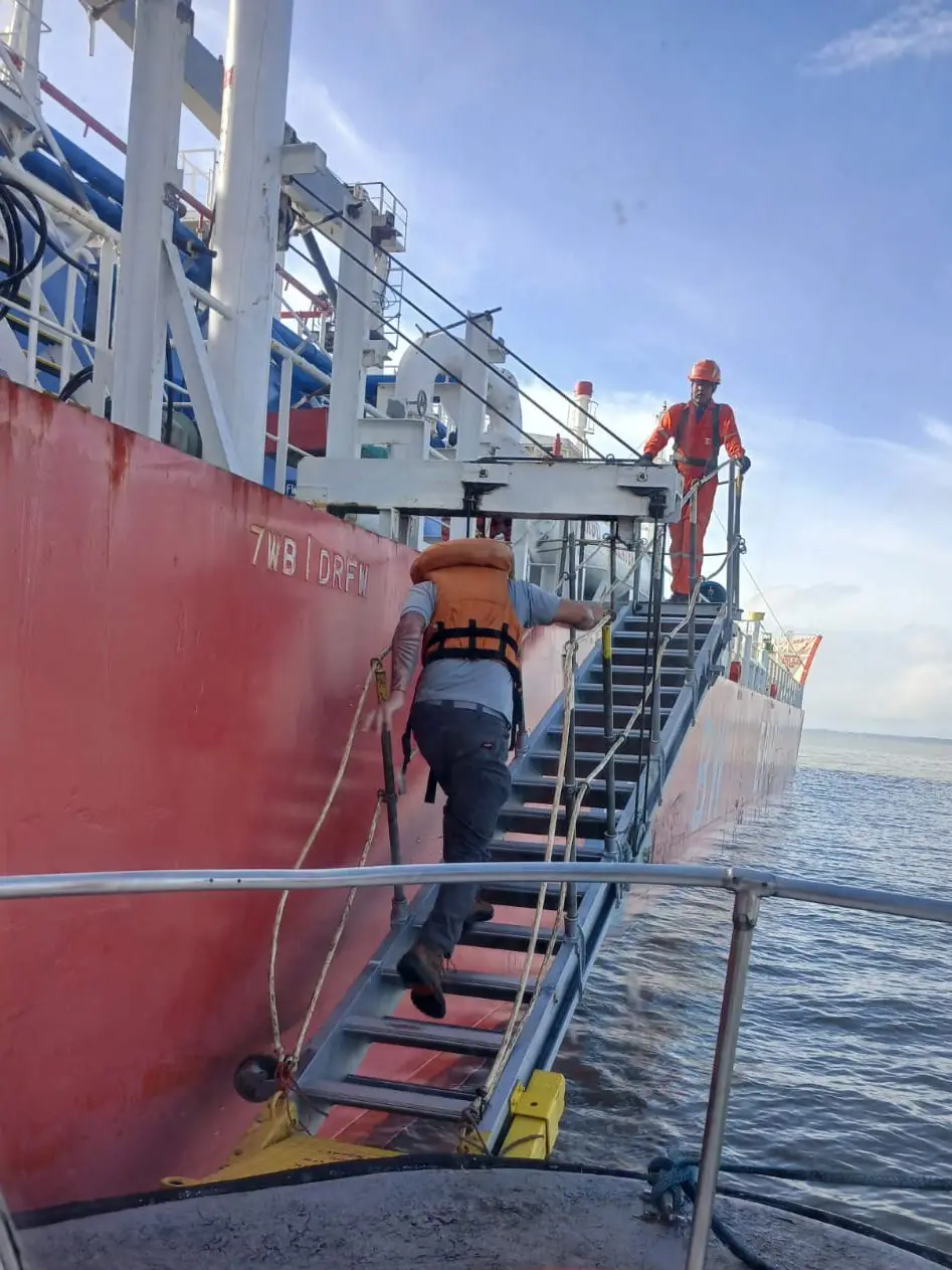 Seafarer in a life jacket climbing a ship's accommodation ladder from a launch, with a crew member waiting at the top