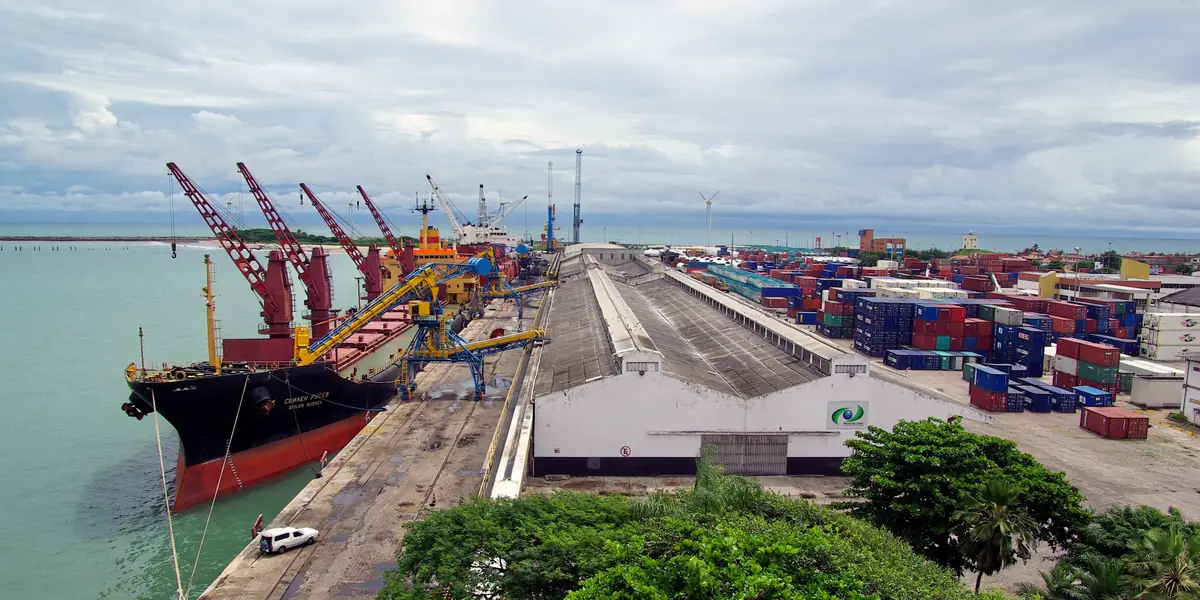 A bulk carrier alongside the commercial quay of the Port of Fortaleza (Mucuripe), with ship unloaders, a long warehouse and the container yard, and wind turbines on the horizon.